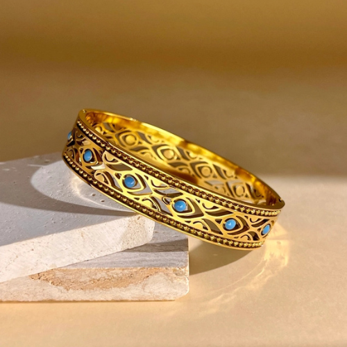 Gesloten goudkleurige stainless steel bangle met blauwe details tentoongesteld op een stuk natuursteen