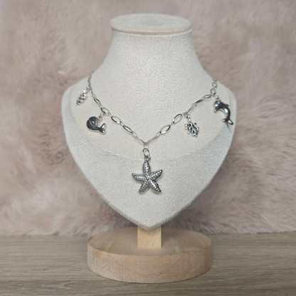 Zilverkleurige bedelketting met zeester, dolfijn, walvis en koraal bedels gepresenteerd op een sieradenstandaard.