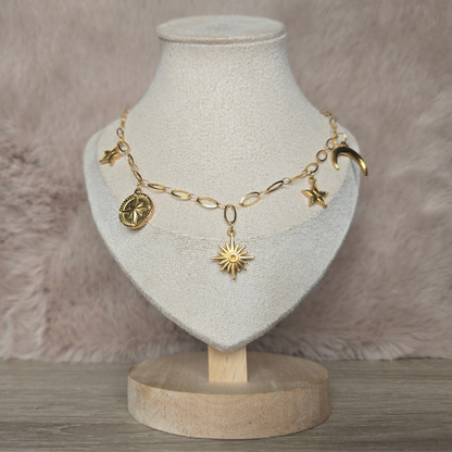 Handgemaakte gouden celestial ketting gepresenteerd op beige sieradenstandaard – met zon, maan en sterren bedels.
